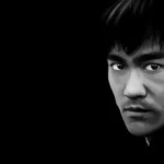Portrait numérique abouti en noir et blanc de Bruce Lee, vue de trois quarts, regard intense et direct, col roulé noir, fond noir uni, traitement en clair-obscur.